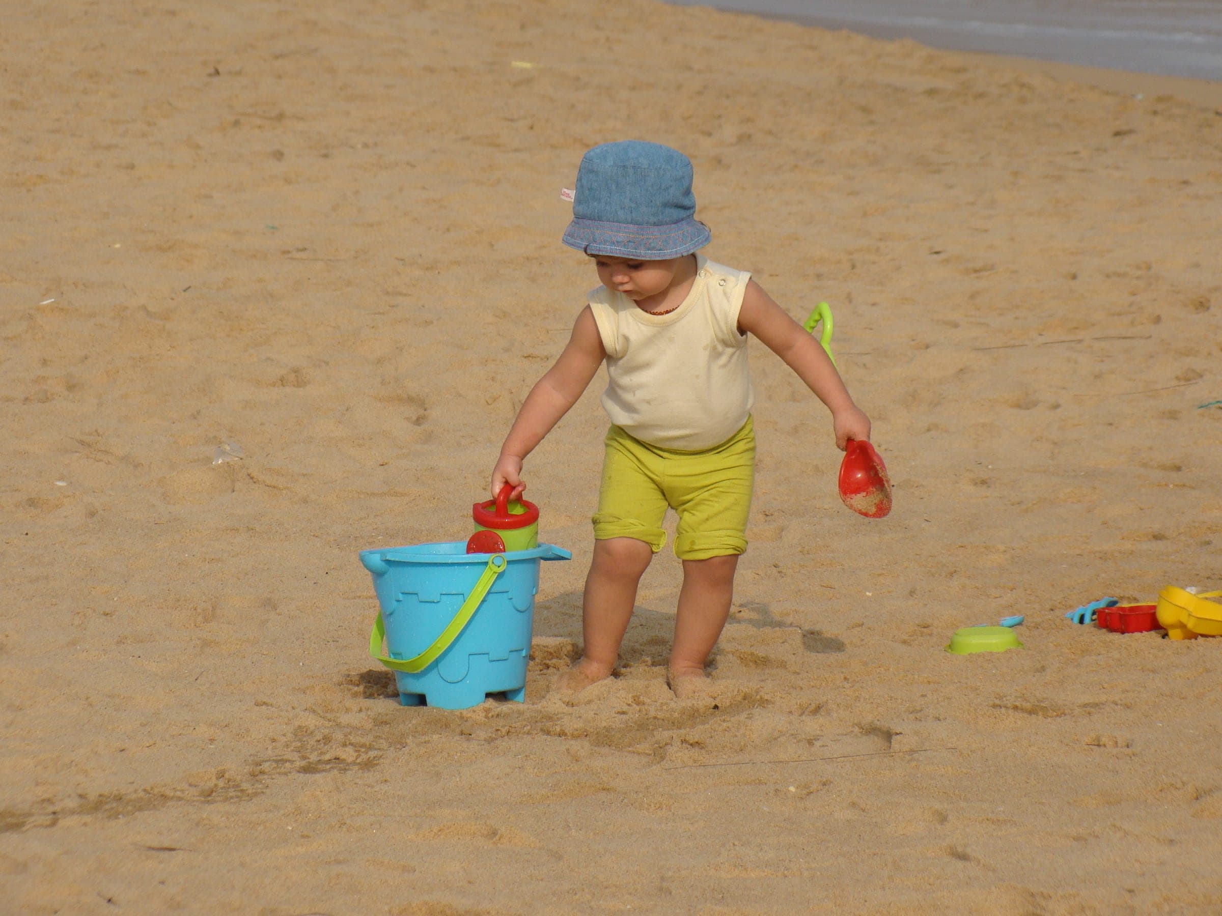 A toddler in a sun hat carrying a blue bucket at the edge of the surf